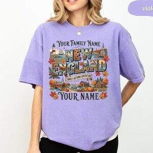 May include: A violet short-sleeve t-shirt featuring a graphic print with the text "YOUR FAMILY NAME", "NEW ENGLAND", "FAMILY AUTUMN ADVENTURE", and "YOUR NAME". The design includes illustrations of a lighthouse, autumn leaves, and buildings.