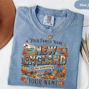 May include: A blue jean coloured t-shirt with a graphic print featuring a lighthouse, autumn leaves, and the text "YOUR FAMILY NAME NEW ENGLAND FAMILY AUTUMN ADVENTURE YOUR NAME". The shirt has a Comfort Colors brand label at the inner neckline.