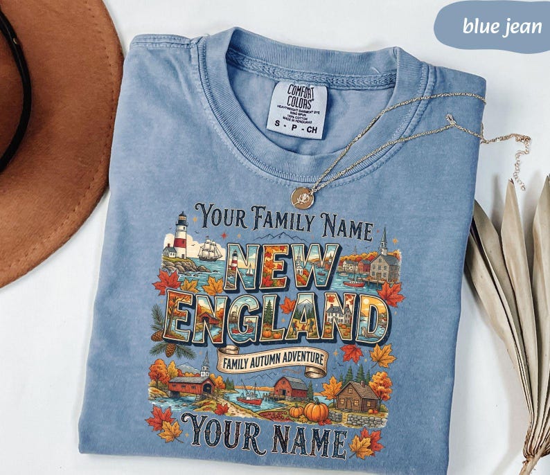 May include: A blue jean coloured t-shirt with a graphic print featuring a lighthouse, autumn leaves, and the text "YOUR FAMILY NAME NEW ENGLAND FAMILY AUTUMN ADVENTURE YOUR NAME". The shirt has a Comfort Colors brand label at the inner neckline.