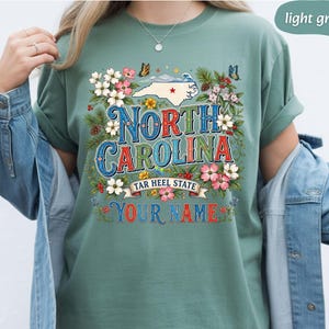 Puede incluir: Camiseta verde claro con un dise&ntilde;o gr&aacute;fico colorido. El dise&ntilde;o incluye las palabras "North Carolina" en una fuente decorativa, un mapa del estado, flores y mariposas. Tambi&eacute;n se incluye el texto "Tar Heel State" y "Your Name".