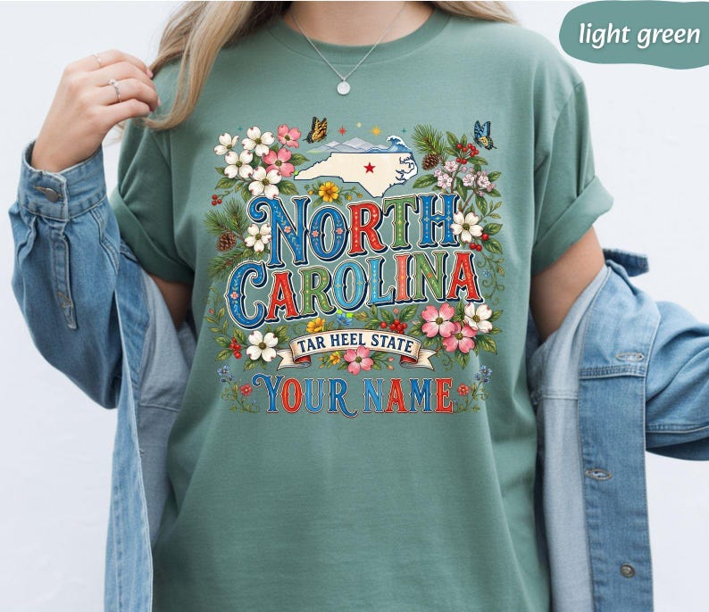 Puede incluir: Camiseta verde claro con un dise&ntilde;o gr&aacute;fico colorido. El dise&ntilde;o incluye las palabras "North Carolina" en una fuente decorativa, un mapa del estado, flores y mariposas. Tambi&eacute;n se incluye el texto "Tar Heel State" y "Your Name".