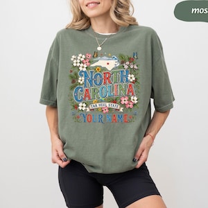 Puede incluir: Una camiseta verde musgo con un gr&aacute;fico colorido de Carolina del Norte, detalles florales y mariposas. El texto "North Carolina Tar Heel State" y "Your Name" tambi&eacute;n se muestran en la camiseta.