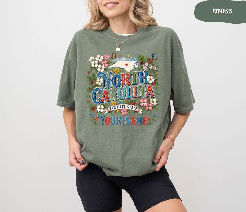 Puede incluir: Una camiseta verde musgo con un gr&aacute;fico colorido de Carolina del Norte, detalles florales y mariposas. El texto "North Carolina Tar Heel State" y "Your Name" tambi&eacute;n se muestran en la camiseta.