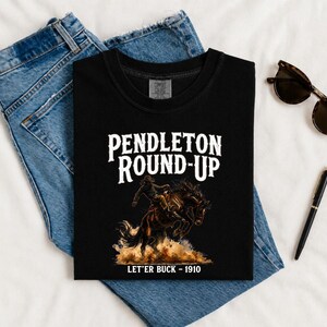 May include: Black t-shirt with the words "Pendleton Round-Up" in white, featuring a graphic of a cowboy riding a bucking bronco. The text "Let'er Buck - 1910" is below the graphic. The shirt is displayed on a pair of blue jeans.