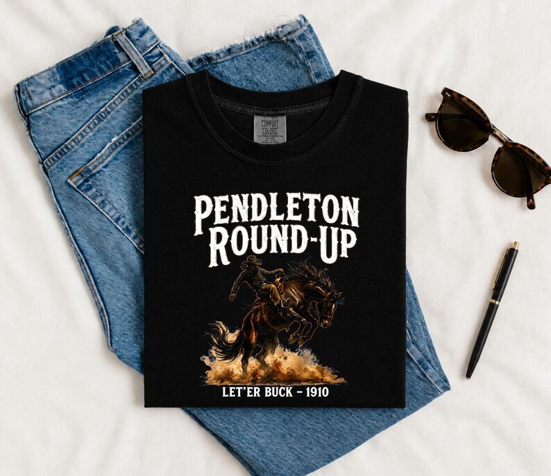 May include: Black t-shirt with the words "Pendleton Round-Up" in white, featuring a graphic of a cowboy riding a bucking bronco. The text "Let'er Buck - 1910" is below the graphic. The shirt is displayed on a pair of blue jeans.