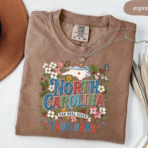 Puede incluir: Camiseta color espresso con un dise&ntilde;o floral y de mariposas alrededor de las palabras "North Carolina". La camiseta presenta el contorno del estado y los textos "Tar Heel State" y "Your Name".