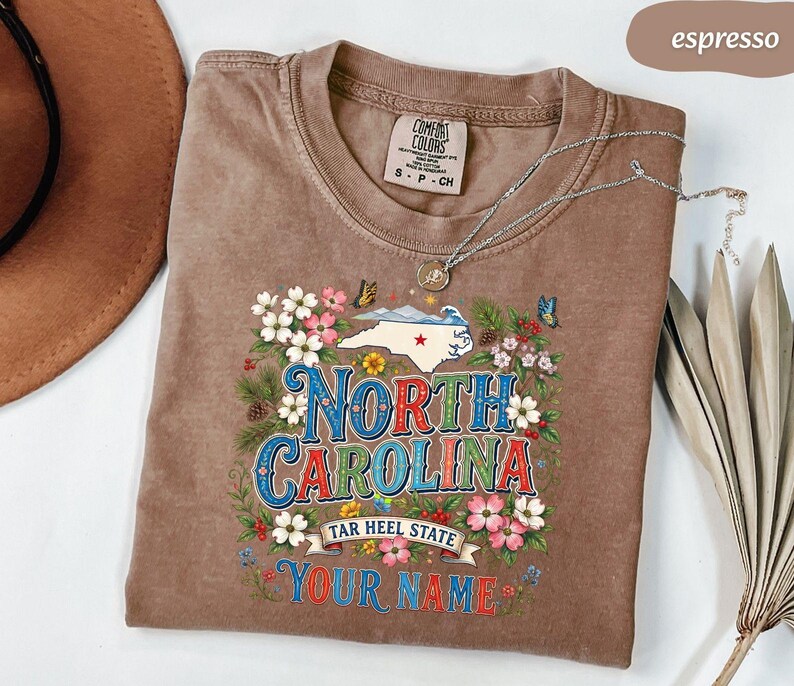 Puede incluir: Camiseta color espresso con un dise&ntilde;o floral y de mariposas alrededor de las palabras "North Carolina". La camiseta presenta el contorno del estado y los textos "Tar Heel State" y "Your Name".