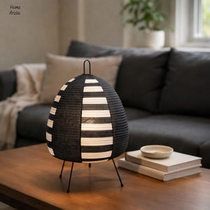 Op de afbeelding: Zwart-witte eivormige tafellamp met een gestreept ontwerp. De lamp heeft een zwart metalen frame met drie poten en een handvat aan de bovenkant. De lamp staat op een houten tafel. De lamp is ongeveer 30 cm hoog.