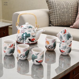 Può includere: Un servizio da tè in ceramica bianca con una teiera e sei tazze. La teiera ha un manico intrecciato ed è decorata con fiori di loto, pesci koi e foglie verdi. Le tazze presentano lo stesso design. Il set è esposto su un tavolo di marmo.