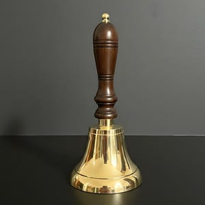 以下が含まれることがあります： 磨かれた真鍮のベルと濃い茶色の木製ハンドル。ベルはフレアベースと丸みを帯びた上部があり、ハンドルの上部には小さな金色のボールが付いています。ベルは手に持って鳴らすように設計されています。