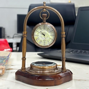 Op de afbeelding: Een vintage messing klok met een zakhorloge-ontwerp, met de tekst "Victoria London 1876". De klok wordt ondersteund door een houten basis en een messing boog. Een kompas is in de basis geïntegreerd. De klok is een decoratief item.