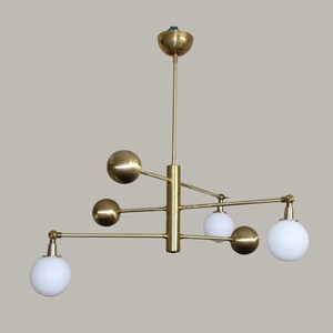 May include: A brass chandelier in a mid-century modern style. The light fitting has a central stem with horizontal arms. It features four brass spheres and three white globe lights, creating a balanced and stylish design.