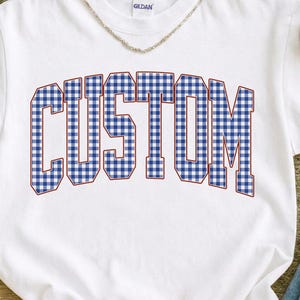 May include: White t-shirt with the word "CUSTOM" in large, bold, blue and white gingham letters. The shirt has a crew neck and short sleeves. The brand name "GILDAN" is visible on the collar.