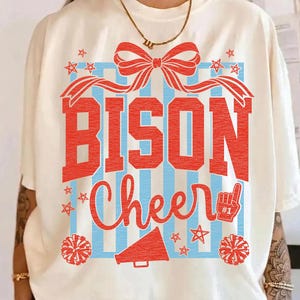 May include: Cream-colored t-shirt with a red and blue graphic. The design features the word "BISON" in large red letters above the word "Cheer" in a script font. Includes stars, a bow, pom-poms, and a megaphone.