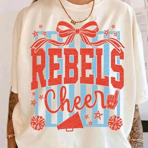 May include: Cream-colored t-shirt with a red and blue graphic. The design includes the words "REBELS Cheer" in a bold font, a bow, stars, a megaphone, and a foam finger. The shirt has short sleeves.