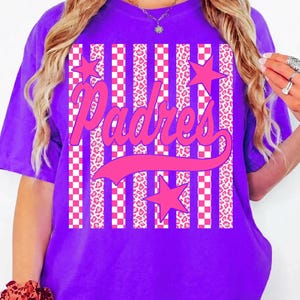 May include: A purple t-shirt featuring the word "Padres" in pink script. The design includes pink stars, checkered patterns, and leopard print stripes. The shirt is a vibrant, eye-catching piece of apparel.