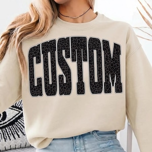 May include: A cream-colored sweatshirt with the word "CUSTOM" in large, block letters across the front. The letters are filled with a black and gray leopard print pattern and outlined in black. The sweatshirt has long sleeves and a crew neck.
