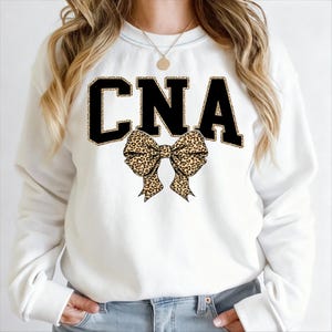 Puede incluir: Sudadera blanca con las letras "CNA" en negro con borde de estampado de leopardo. Un lazo con estampado de leopardo está centrado debajo de las letras. La sudadera es de cuello redondo.