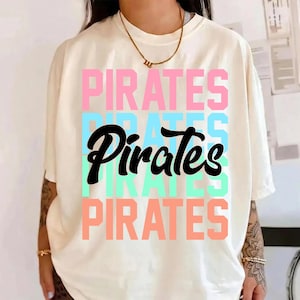 May include: Cream-colored t-shirt with the word "PIRATES" in a stacked design. The word is in pink, blue, and green, with a black cursive "Pirates" in the center. The shirt has short sleeves.