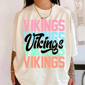 Può includere: T-shirt color crema con la scritta "VIKINGS" in lettere rosa, blu e corallo, e un "Vikings" nero in corsivo al centro. La maglietta ha maniche corte.