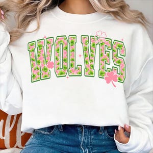 Peut inclure: Sweat-shirt blanc avec le mot "WOLVES" en grandes lettres bordées de vert, remplies d'un motif floral rose et de trèfles verts. Des accents de trèfles roses et des étoiles sont également présents.