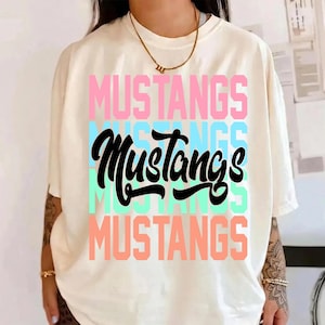 May include: Cream-colored t-shirt with the word "MUSTANGS" in pink, blue, black, and green. The word is stacked in a repeating pattern. The shirt has short sleeves and a crew neck.