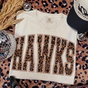 May include: A cream-colored short-sleeve t-shirt featuring the word HAWKS in large, block-style letters. The letters have a brown and black leopard print pattern with a thin white outline. The shirt has a crew neckline and a tag at the inner collar.