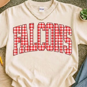 May include: Cream-colored t-shirt with the word "FALCONS" in large, red and white gingham letters. The shirt has a crew neck and short sleeves. A silver chain necklace is visible at the neckline.