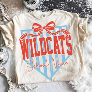 以下が含まれることがあります： 赤色のリボンと、赤いブロック体の「WILDCATS」の文字が入ったクリーム色のTシャツ。テキストは、水色のストライプのシールドの形に対して設定されています。「WILDCATS」の下には、赤いスクリプトで「School Name」というテキストがあります。