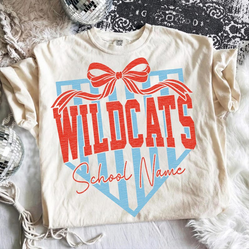 以下が含まれることがあります： 赤色のリボンと、赤いブロック体の「WILDCATS」の文字が入ったクリーム色のTシャツ。テキストは、水色のストライプのシールドの形に対して設定されています。「WILDCATS」の下には、赤いスクリプトで「School Name」というテキストがあります。