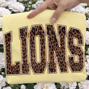 May include: A pale yellow t-shirt featuring the word LIONS in large, block-style varsity lettering. The letters have a brown and black leopard print pattern with a distressed, weathered texture and a thin brown outline surrounding each character.