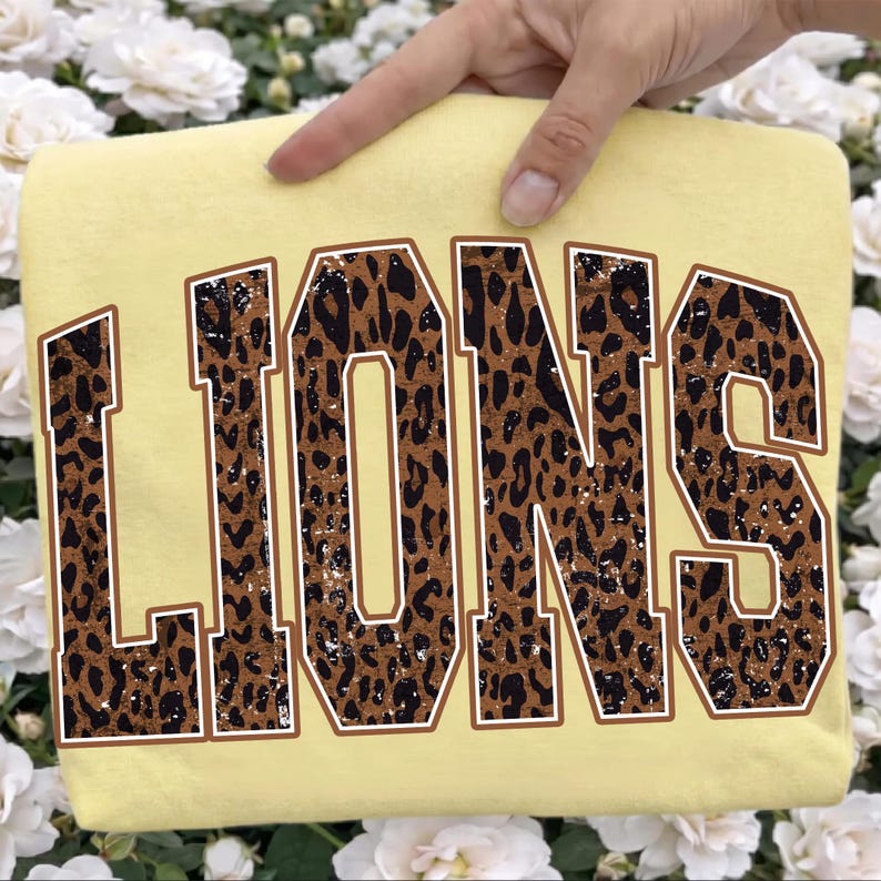 May include: A pale yellow t-shirt featuring the word LIONS in large, block-style varsity lettering. The letters have a brown and black leopard print pattern with a distressed, weathered texture and a thin brown outline surrounding each character.
