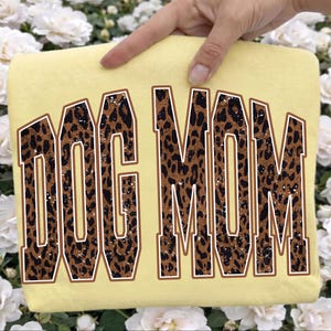 May include: A pale yellow t-shirt featuring the text DOG MOM in a large, arched, collegiate-style font. The letters have a brown and black leopard print pattern with a distressed, weathered texture and a thin brown outline around each letter.