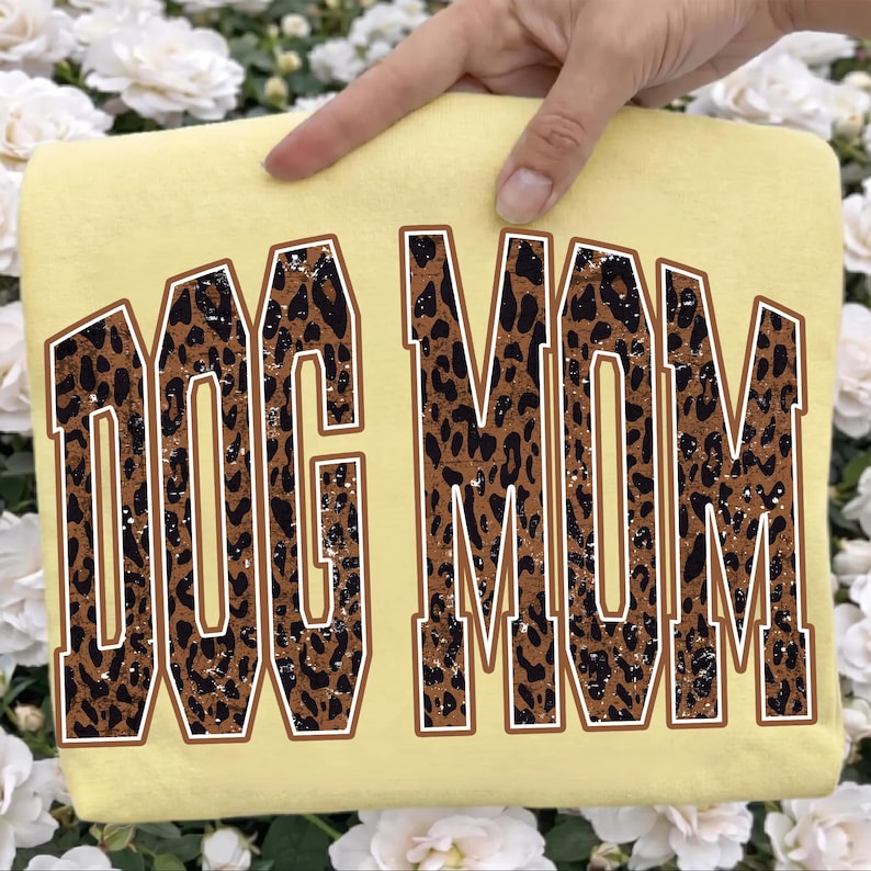 May include: A pale yellow t-shirt featuring the text DOG MOM in a large, arched, collegiate-style font. The letters have a brown and black leopard print pattern with a distressed, weathered texture and a thin brown outline around each letter.
