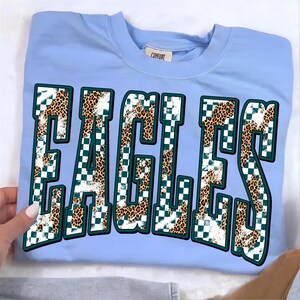 May include: A light blue crewneck t-shirt featuring the word EAGLES in large, block-style letters. The letters have a pattern consisting of a white and dark green checkerboard design overlaid with a brown and black leopard print texture.