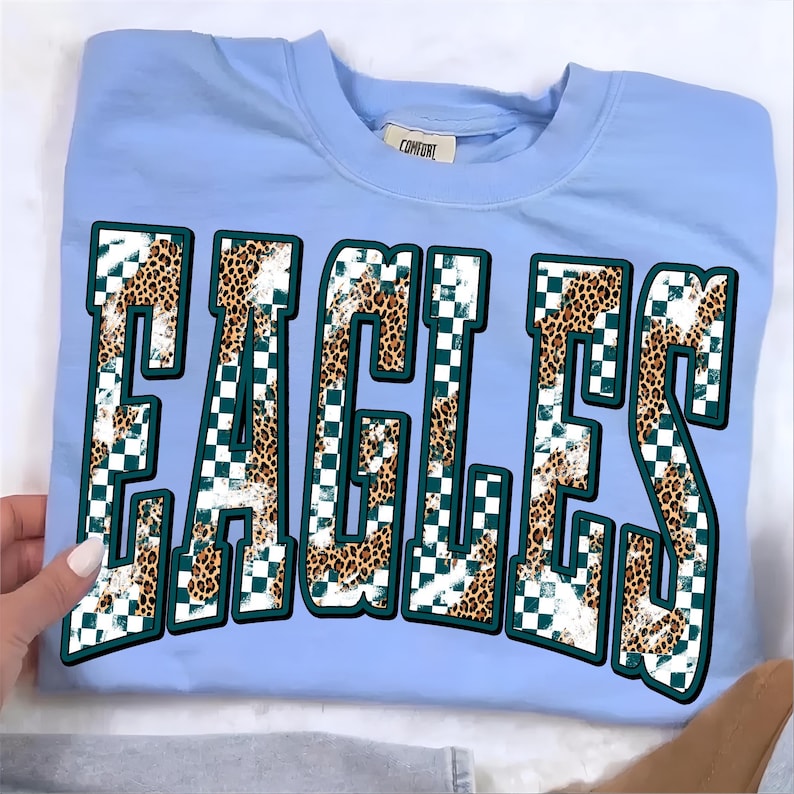May include: A light blue crewneck t-shirt featuring the word EAGLES in large, block-style letters. The letters have a pattern consisting of a white and dark green checkerboard design overlaid with a brown and black leopard print texture.