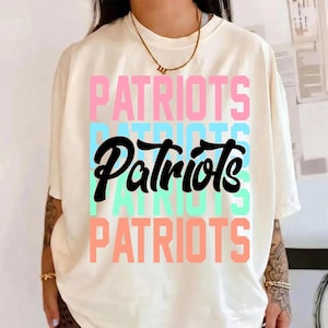 May include: Cream-colored t-shirt with the word "PATRIOTS" stacked vertically in pink, light blue, and coral. The word "Patriots" is also written in a bold, black script. The shirt has short sleeves.