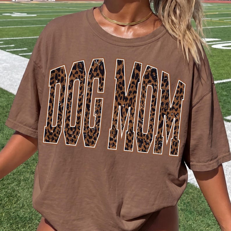May include: A brown short-sleeved t-shirt featuring the text DOG MOM in large, block-style letters. The letters have a brown and black leopard print pattern with a white outline. The shirt has a crew neckline and a relaxed, loose-fitting silhouette.