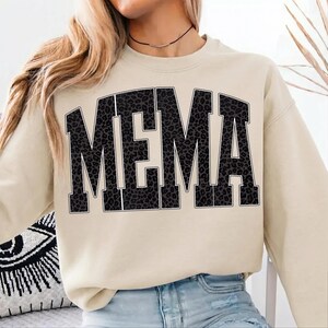 May include: A beige sweatshirt with the word "MEMA" in large, black, leopard print letters across the front. The sweatshirt has long sleeves and a crew neck. The model is wearing blue jeans.