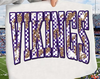 May include: A white t-shirt featuring the word VIKINGS in large, bold, purple-outlined letters. The interior of each letter is filled with a pattern of purple and white checkerboard squares mixed with a brown and black leopard print design.
