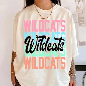 Peut inclure: T-shirt crème avec le mot "WILDCATS" dans un design superposé. Les couches supérieure et inférieure sont roses, la couche du milieu est bleue et le texte central est noir. Un collier doré est visible.