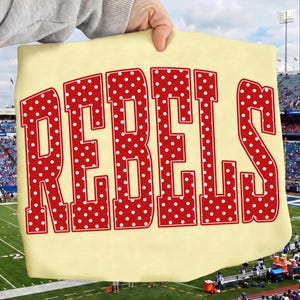 May include: A light yellow t-shirt featuring the word REBELS in large, bold, red block letters. Each letter is filled with a pattern of small white polka dots and outlined with a thin red border, creating a distinct graphic design on the fabric.