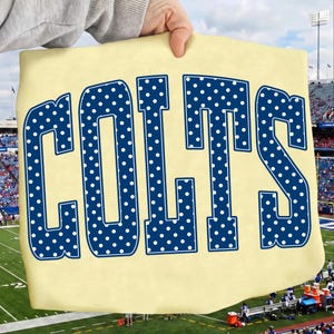 Puede incluir: Una camiseta de color amarillo claro con la palabra COLTS en letras mayúsculas grandes y de color azul oscuro. Cada letra está decorada con un patrón repetitivo de pequeños puntos blancos. El texto está centrado en la prenda.