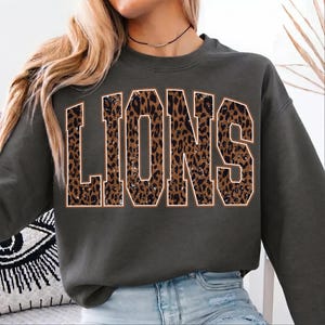 May include: A dark gray crewneck sweatshirt featuring the word LIONS in large, block-style letters. The letters have a brown and black leopard print pattern with a thin white outline, centered on the front of the garment.