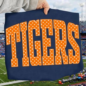 Puede incluir: Una camiseta de color azul marino oscuro con la palabra TIGERS en letras mayúsculas grandes y naranjas. Cada letra está rellena con un patrón repetitivo de pequeños puntos blancos. El diseño está centrado en la parte frontal.