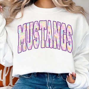 May include: White sweatshirt with the word "MUSTANGS" in large, purple, block letters. The letters are filled with a pastel pattern of bunnies and flowers. The sweatshirt is a casual style.