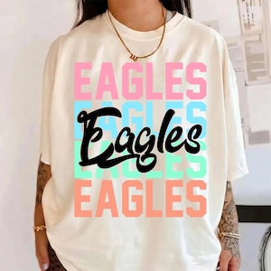 May include: Cream-colored t-shirt with the word "EAGLES" stacked in pink, blue, and green. The word "Eagles" is written in a cursive font in black. A gold necklace is visible.