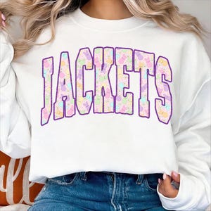 May include: White sweatshirt with the word "JACKETS" in large, purple outlined letters. The letters are filled with a pattern of pastel-colored bunnies and flowers. The sweatshirt is paired with blue jeans.
