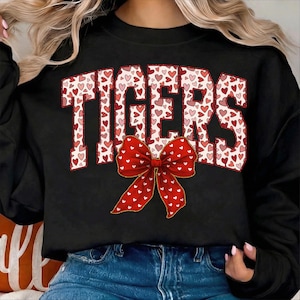 May include: Black sweatshirt with the word "TIGERS" in a red and pink heart pattern. A large red bow with white hearts is centered below the word. The sweatshirt is a casual style.
