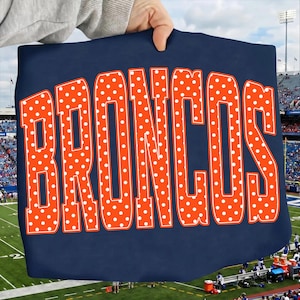 Puede incluir: Una camiseta de color azul marino oscuro con la palabra BRONCOS en letras grandes, negritas y de color naranja. Cada letra está rellena con un patrón repetitivo de pequeños puntos blancos y delineada con un borde blanco fino, creando un diseño gráfico de alto contraste.